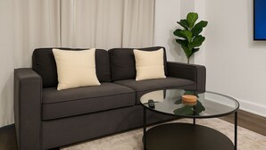 A modern living room with a dark grey sofa, two cream-colored pillows, a glass coffee table with a wooden coaster, and a green plant. - Chic Studio in Historic Hyde Park – Walk to Campus (Chicago)