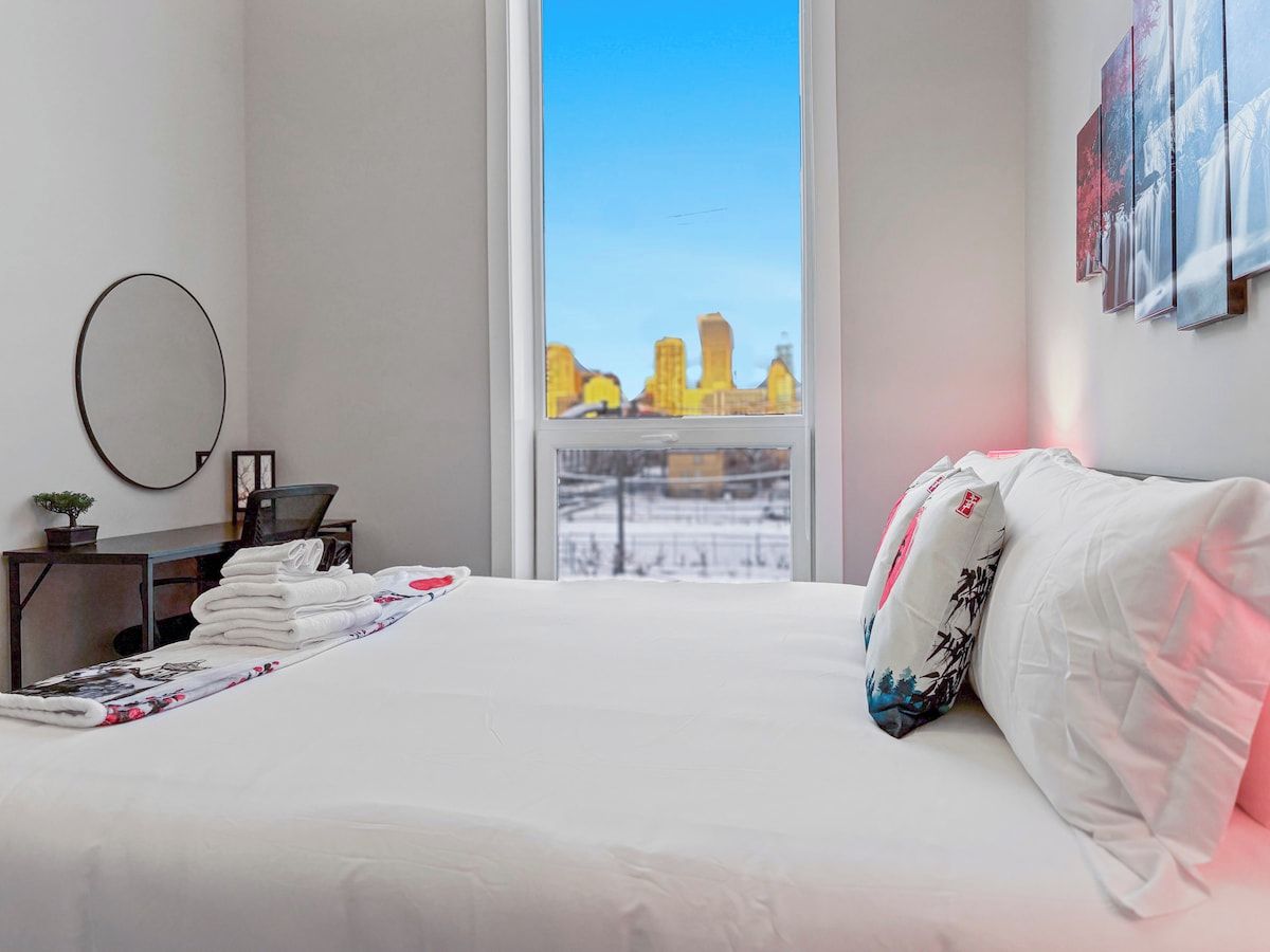 Quarto de hotel com uma cama, escrivaninha com cadeira, espelho redondo e janela com vista para a cidade.
