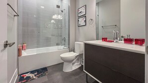 Un baño moderno con cabina de ducha de vidrio, inodoro blanco, tocador oscuro con espejo y una alfombra pequeña en el suelo.