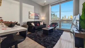 Una sala moderna con un sofá, una mesa de centro y un comedor con mesa y sillas. Hay un ventanal con vistas a un paisaje urbano.