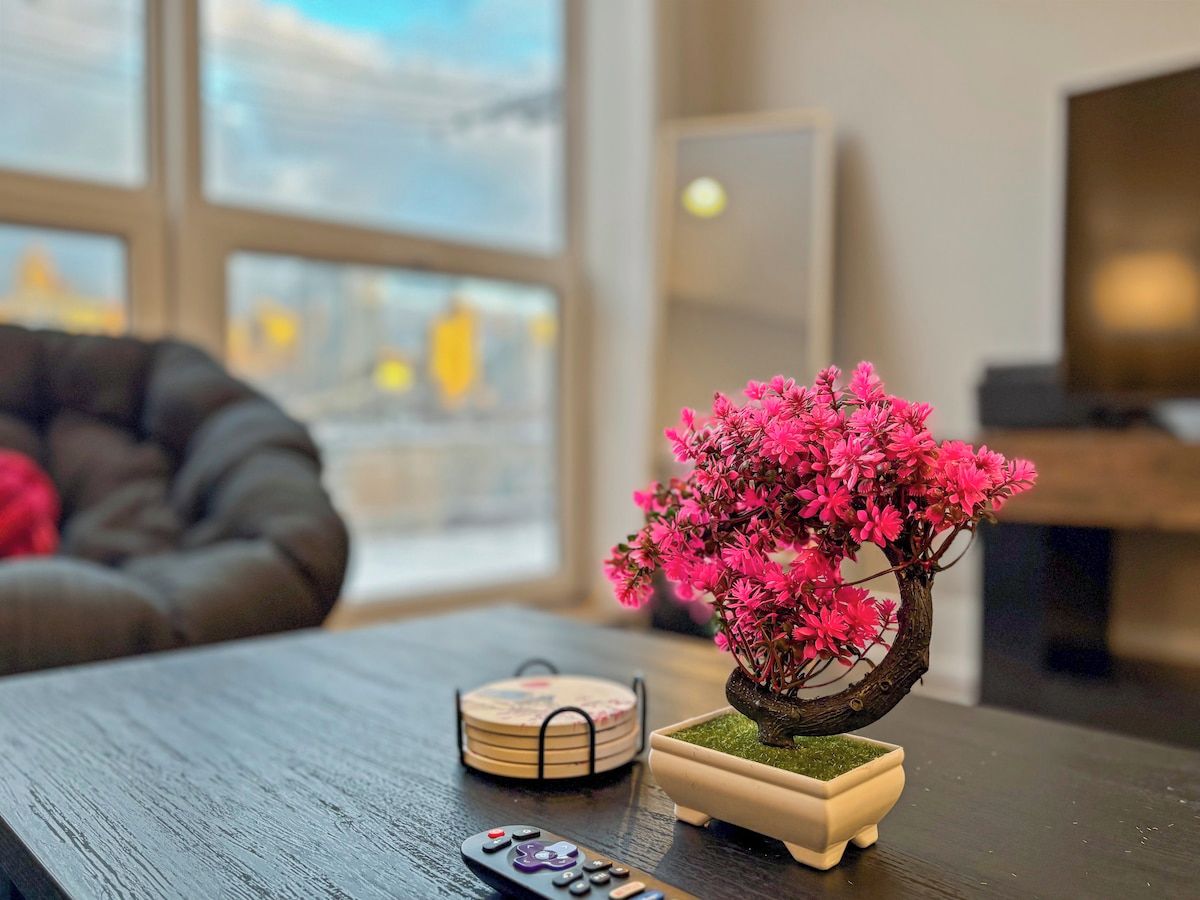 Een bonsaiboom met roze bloemen op een tafel, een afstandsbediening en een stapel onderzetters.