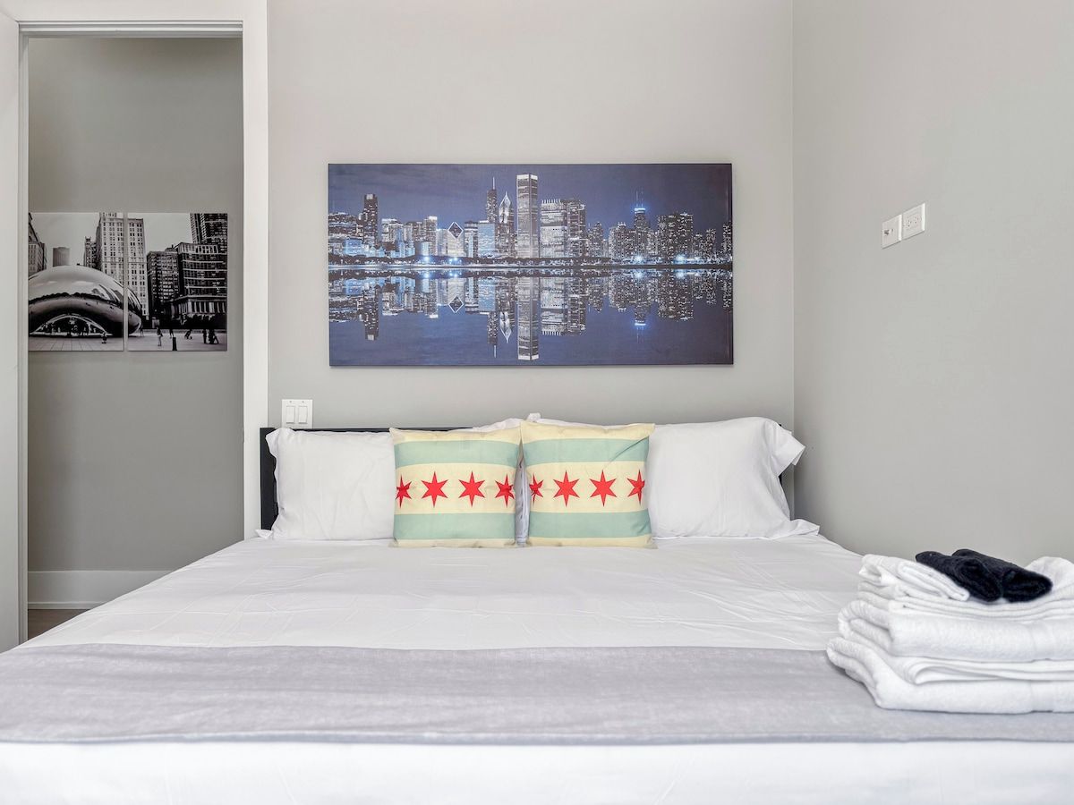A hotel room with a bed, pillows, a cityscape painting, and a neatly folded towel on the side.