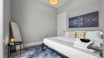 Une chambre d’hôtel avec un lit, une œuvre d’art représentant un paysage urbain, une table de chevet avec une lampe et un tapis bleu à motifs.