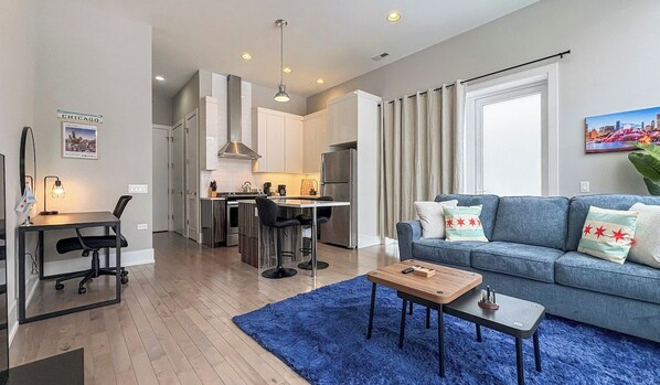Een moderne woonkamer met een blauw vloerkleed, een bank met decoratieve kussens, een houten salontafel en een bureau met een stoel.