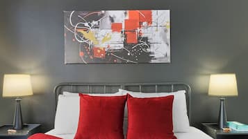 Habitación de hotel con cama, dos lámparas de noche, una manta roja y un cuadro abstracto colgado en la pared.