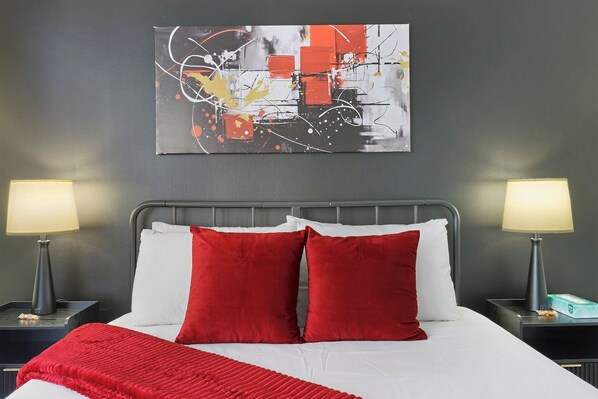 A hotel room with a bed, two bedside lamps, a red throw blanket, and a wall-mounted abstract painting.