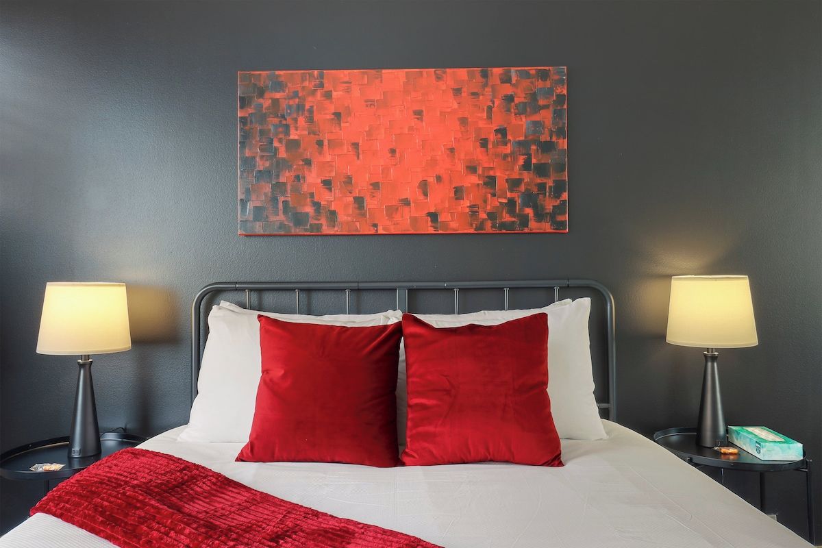 Habitación de hotel con una cama, dos lámparas de noche, una colcha roja y un cuadro abstracto rojo y negro en la pared.