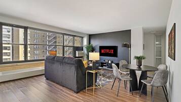 Una sala de estar moderna con sofá, una mesa de comedor con sillas, televisor y un ventanal con vista a edificios.