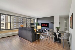 A modern living room with a sofa, a dining table with chairs, a TV, and a large window with a view of buildings.