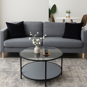 Un canapé gris avec des coussins noirs, une table basse ronde en verre ornée d’un vase et de fleurs, et une petite table blanche avec une plante en arrière-plan.