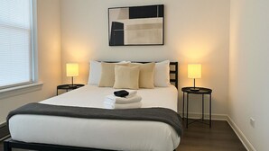 A neatly made bed with a white duvet and a dark throw blanket, flanked by two bedside tables with lamps, a modern abstract painting above, and a window with blinds on the left. - Hyde Park 1BR w/ Charm (Chicago)