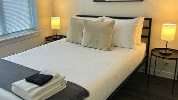 Una cama bien hecha con almohadas y una colcha, dos lámparas de noche, una ventana con persianas y una obra de arte abstracta enmarcada en la pared.