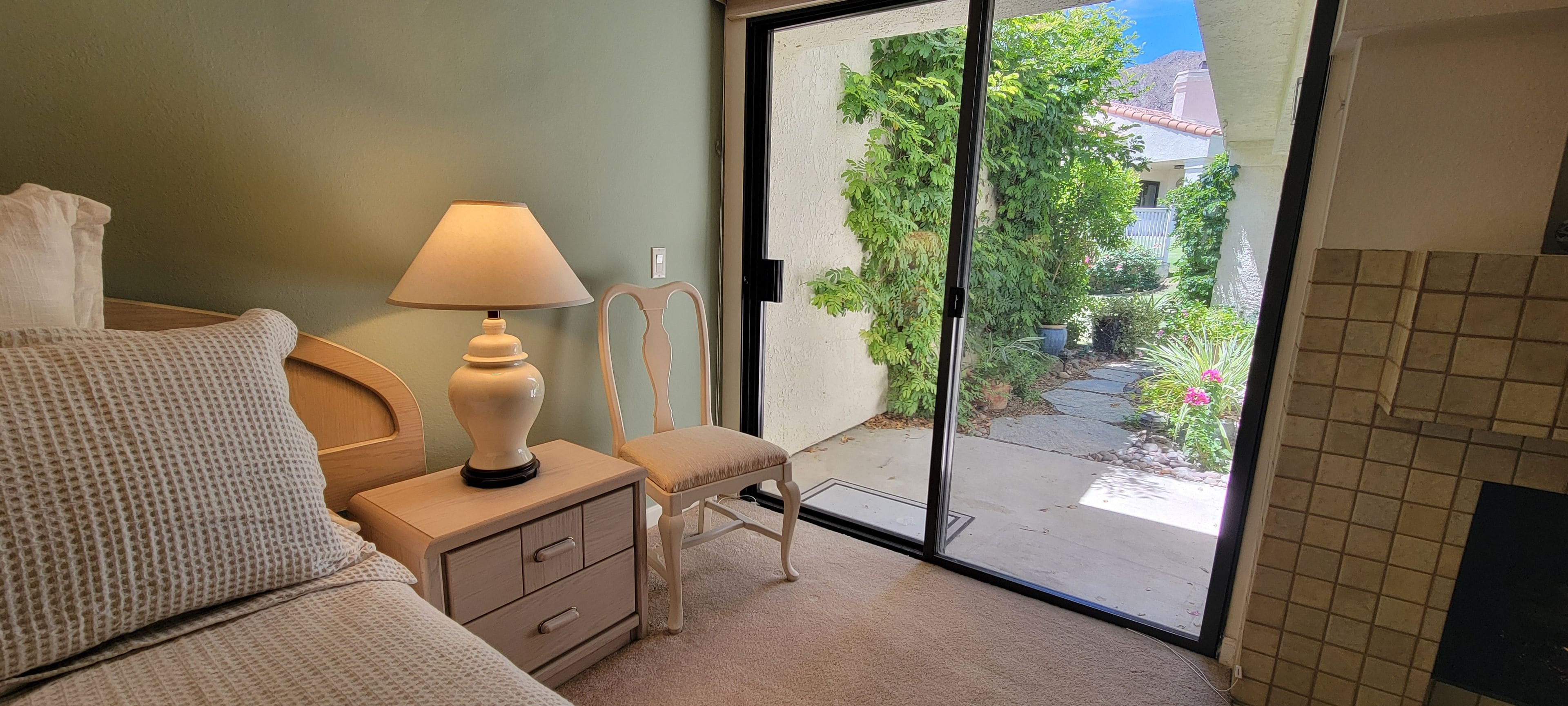 A bedroom with a bed, a nightstand, a lamp, a chair, and a view of a garden through a glass door.