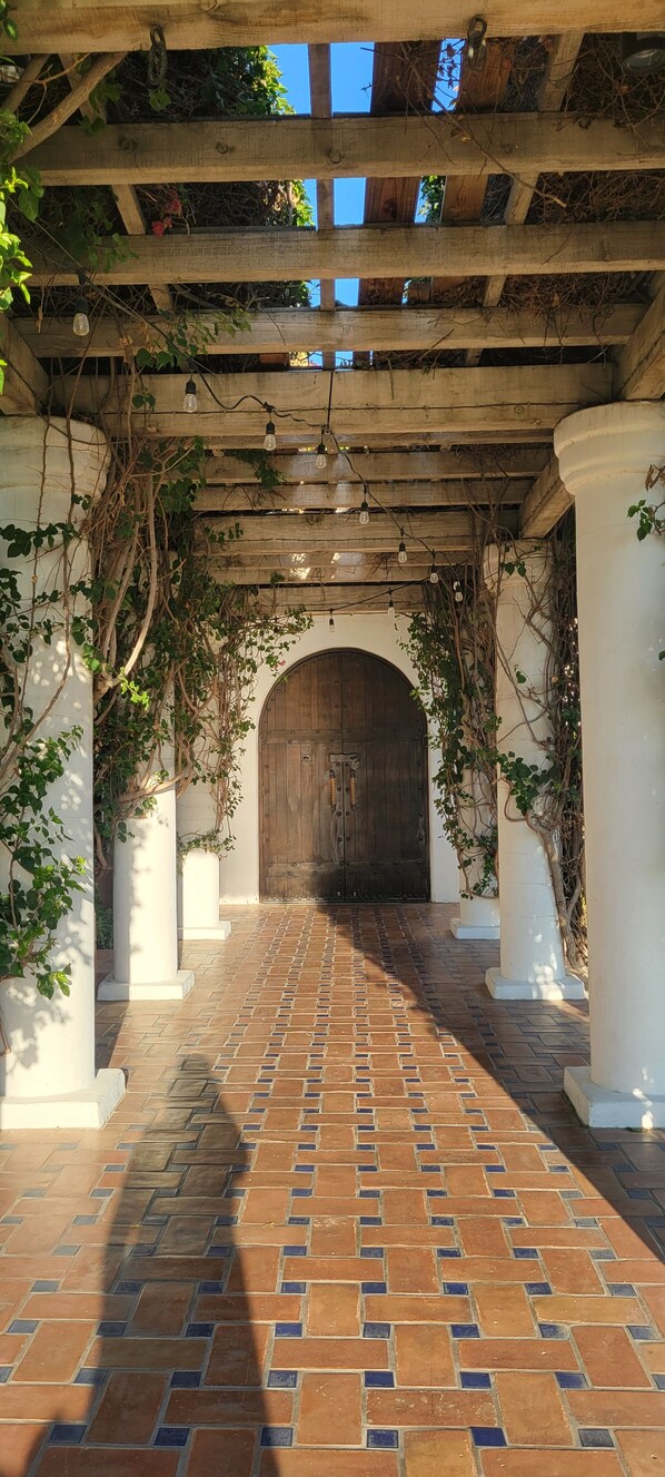 A covered walkway with a brick floor, white columns, and a wooden door at the end.