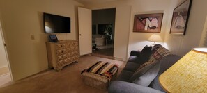 A living room with a flat-screen TV, a wooden dresser, a patterned ottoman, a sofa, and a lamp.