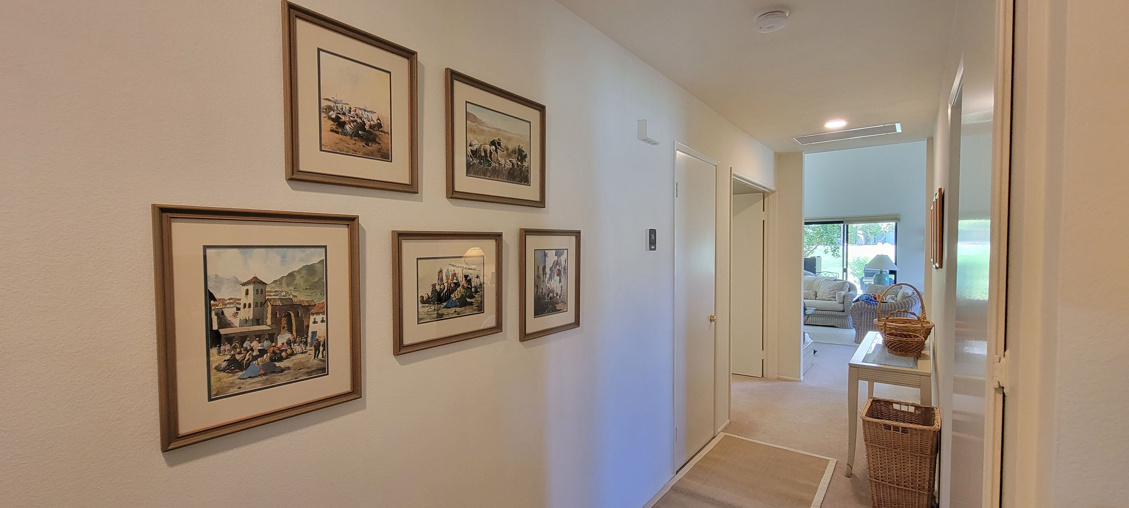 A hallway with framed pictures on the wall, a door leading to a room, and a small table with baskets.