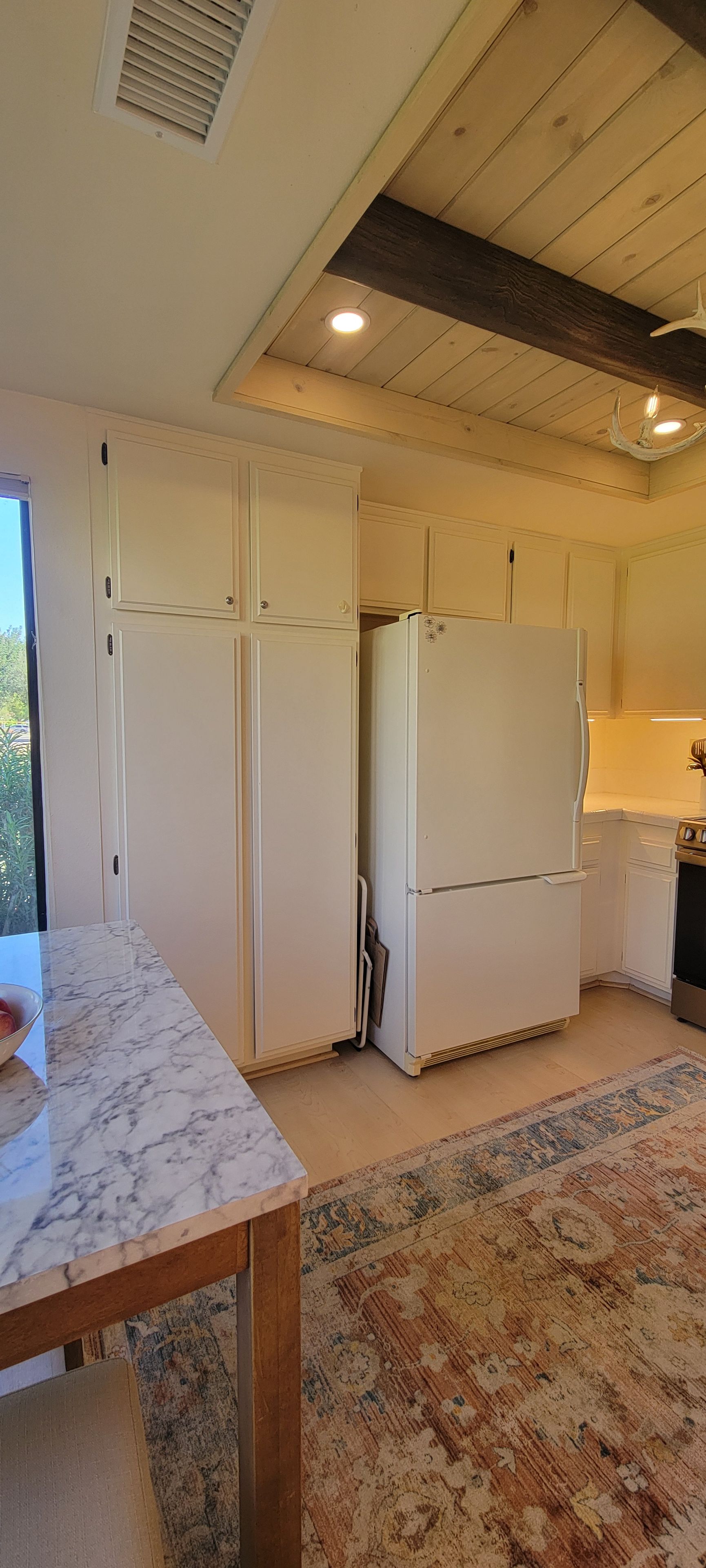 A kitchen with white cabinets, a marble countertop, a patterned rug, and a ceiling with wooden beams.