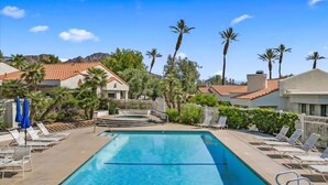A rectangular swimming pool with a clear blue water surface, surrounded by lounge chairs and umbrellas, with a mountainous backdrop and residential buildings.