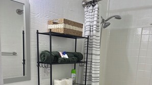 Ein Badezimmer mit weißem Waschbecken, WC und Dusche. Ein schwarzes Regal zeigt Handtücher und Pflegeprodukte.