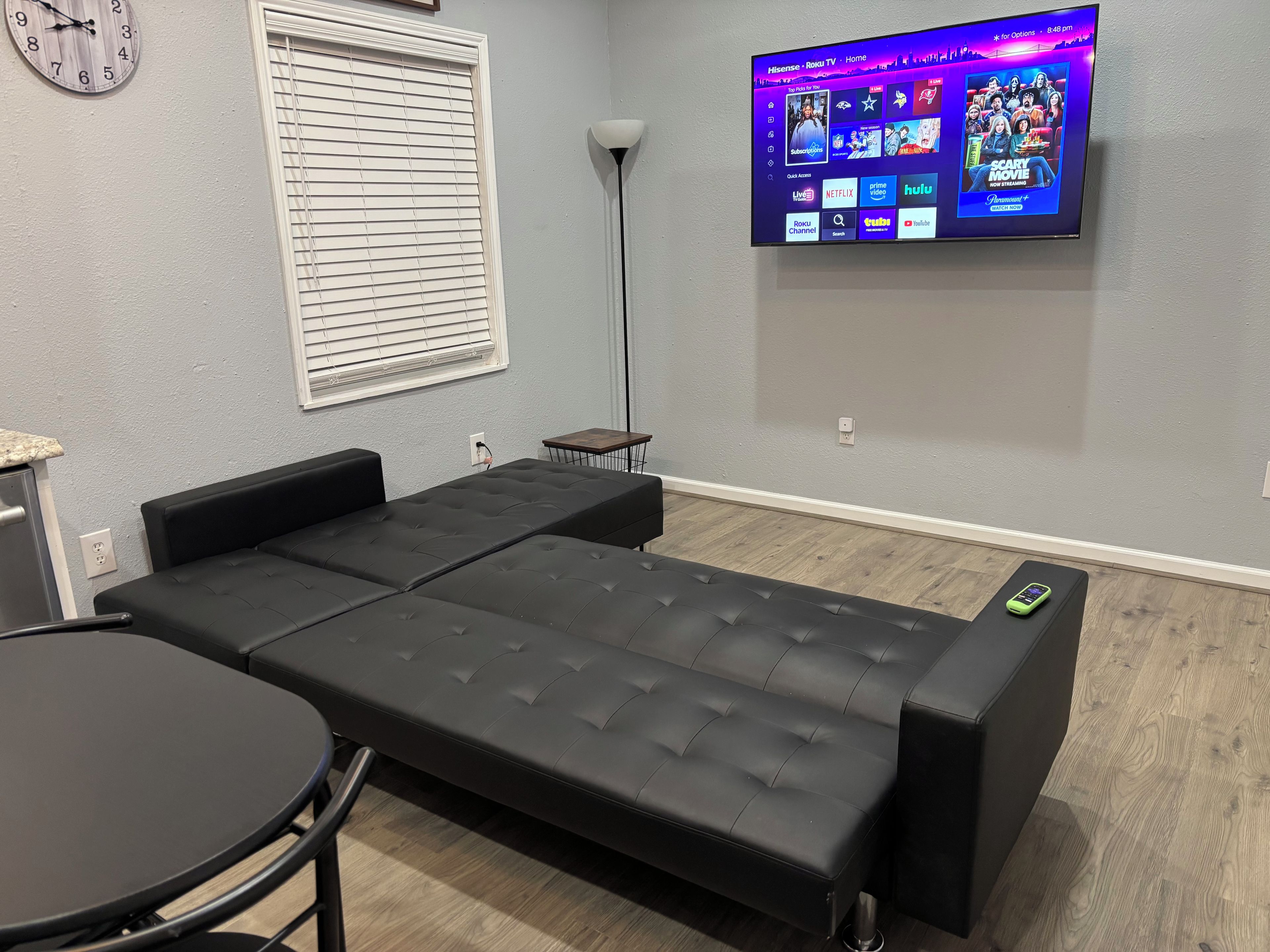 Una habitación con un sofá negro, una mesita redonda pequeña, un televisor empotrado que muestra servicios de streaming y un reloj en la pared.