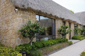 A stone building with a thatched roof, large windows, and small trees in the foreground.