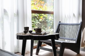 A table with a teapot and cups, a chair, and a view of greenery through a window.