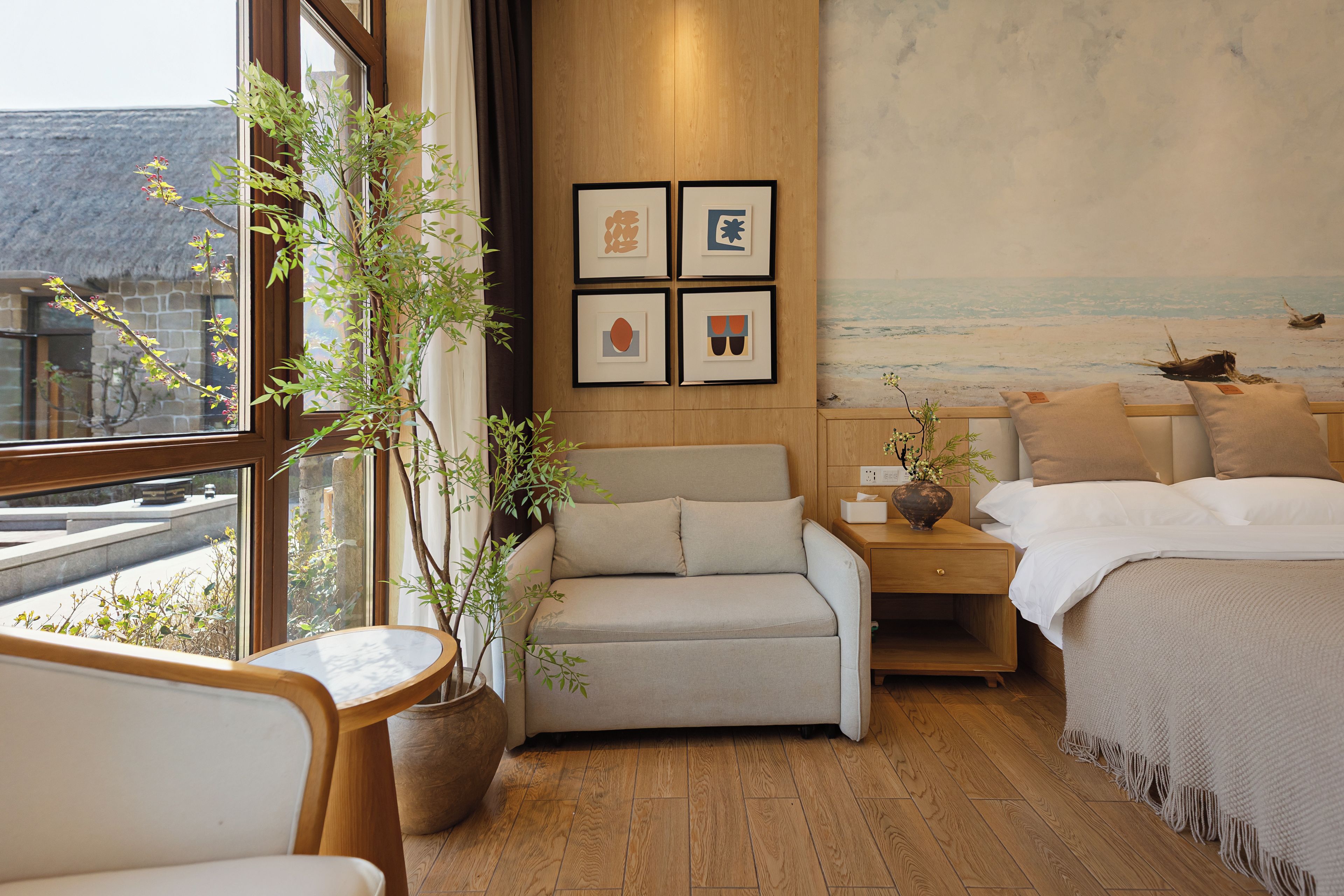 Una habitación de hotel con cama, sofá, mesita de noche, planta en maceta y un cuadro en la pared.