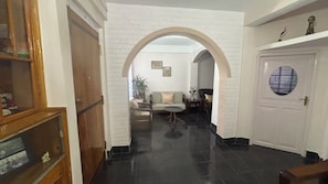 A hallway with a white brick wall, a wooden door, and a round window. There is a sofa and a table with a vase in the background.