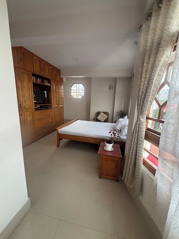 A bedroom with a bed, a small table with a plant, wooden cabinets, and a window with curtains.