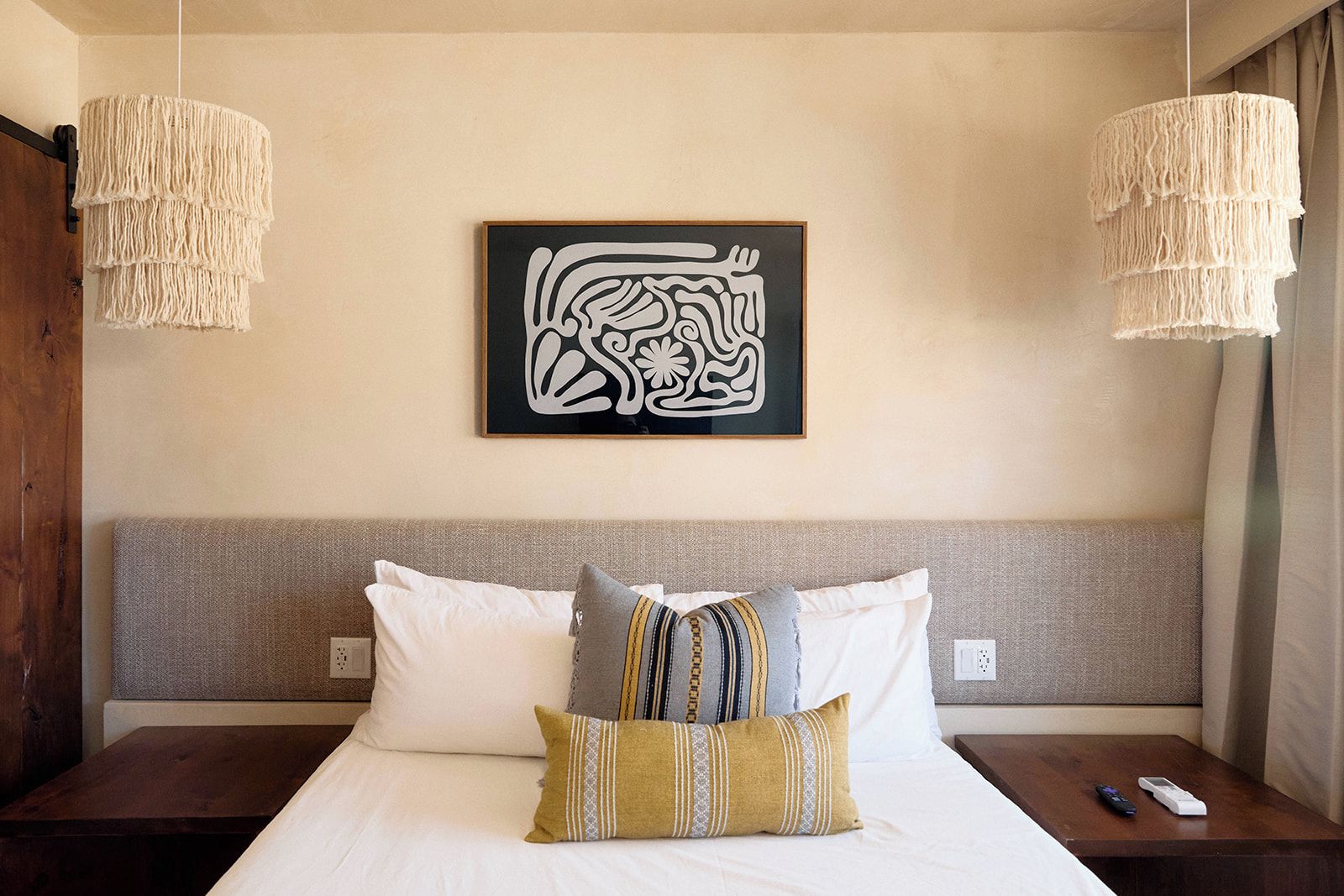 A hotel room with a bed, two bedside tables, a wall-mounted artwork, and two hanging light fixtures.
