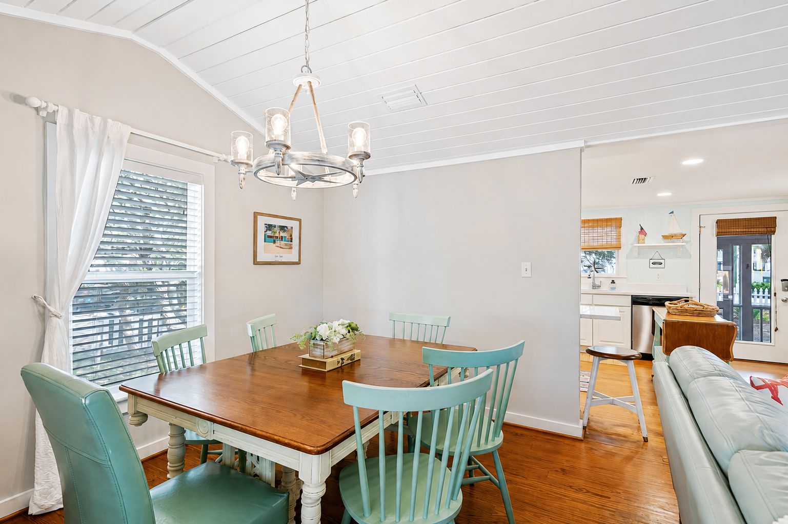 A dining area with a wooden table and teal chairs, a chandelier, and a view of the kitchen through an open door.