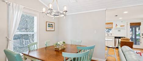 A dining area with a wooden table and teal chairs, a chandelier, and a view of the kitchen through an open door.