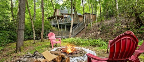 A cabin with a deck and stairs leading to a second level, surrounded by a forest.