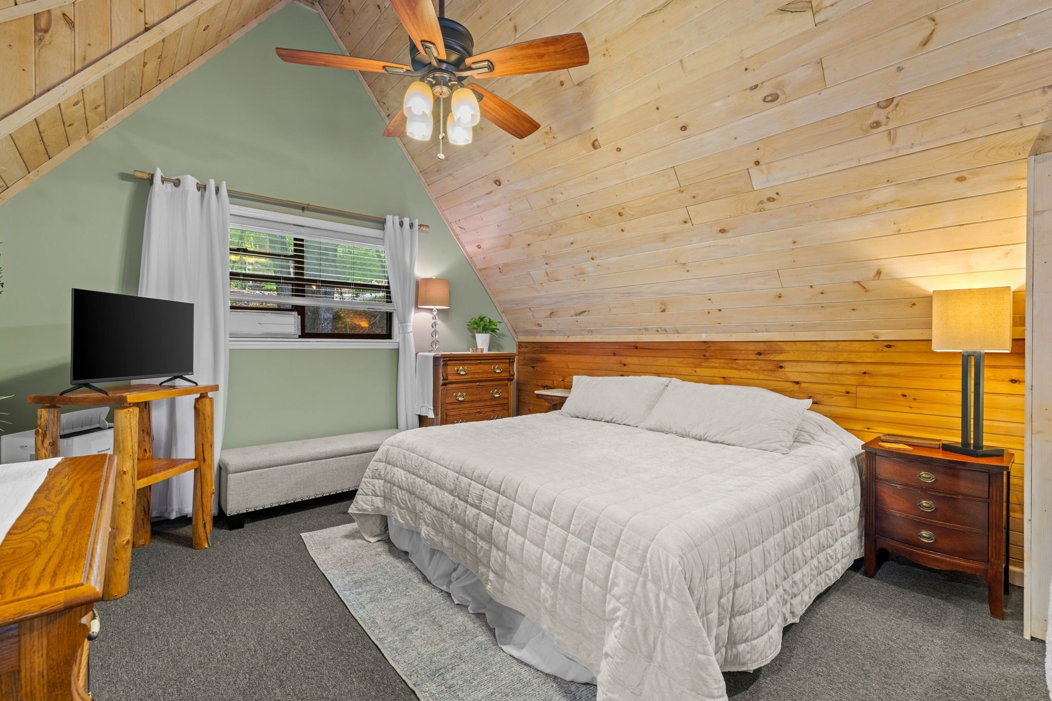 A bedroom with a bed, a desk, a television, a ceiling fan, and a window with curtains.