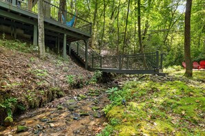 A wooden bridge over a stream in a forested area with a building on the left. - Serenity by the Creek (Robbinsville)