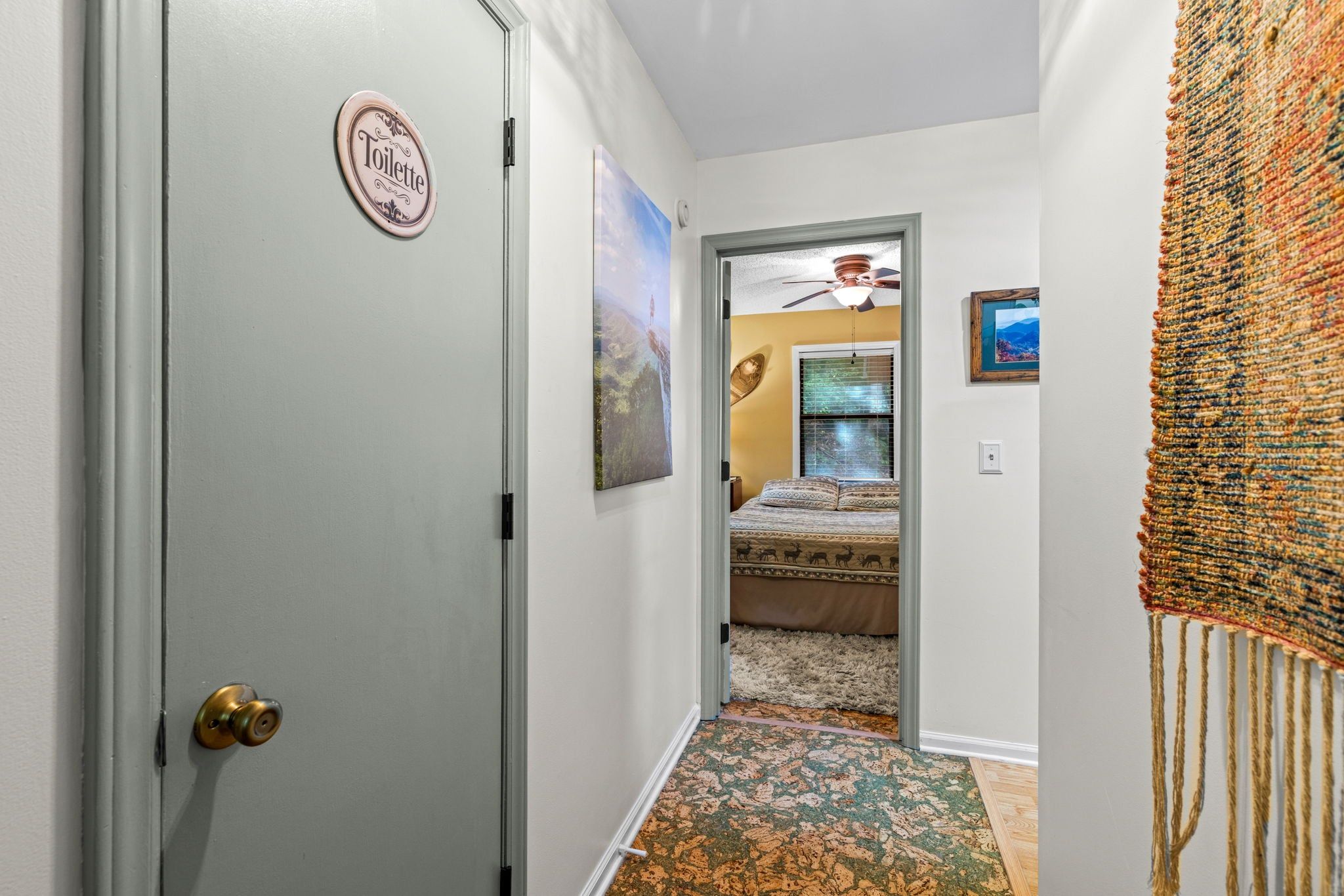 A hallway leading to a bedroom with a bed, a ceiling fan, and a door with a 'Toilette' sign.