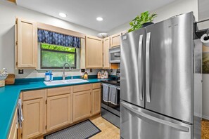 A modern kitchen with wooden cabinets, a stainless steel refrigerator, and a window with a view of greenery. - Serenity by the Creek (Robbinsville)