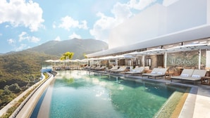 Piscina infinita no terraço com espreguiçadeiras e guarda-sóis, com vista para uma paisagem montanhosa.