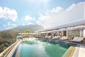 Piscina infinita en la azotea con sillas de descanso y sombrillas, con vistas a un paisaje montañoso.