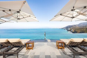 A poolside area with lounge chairs and umbrellas, overlooking a clear blue sea and a hilly landscape.