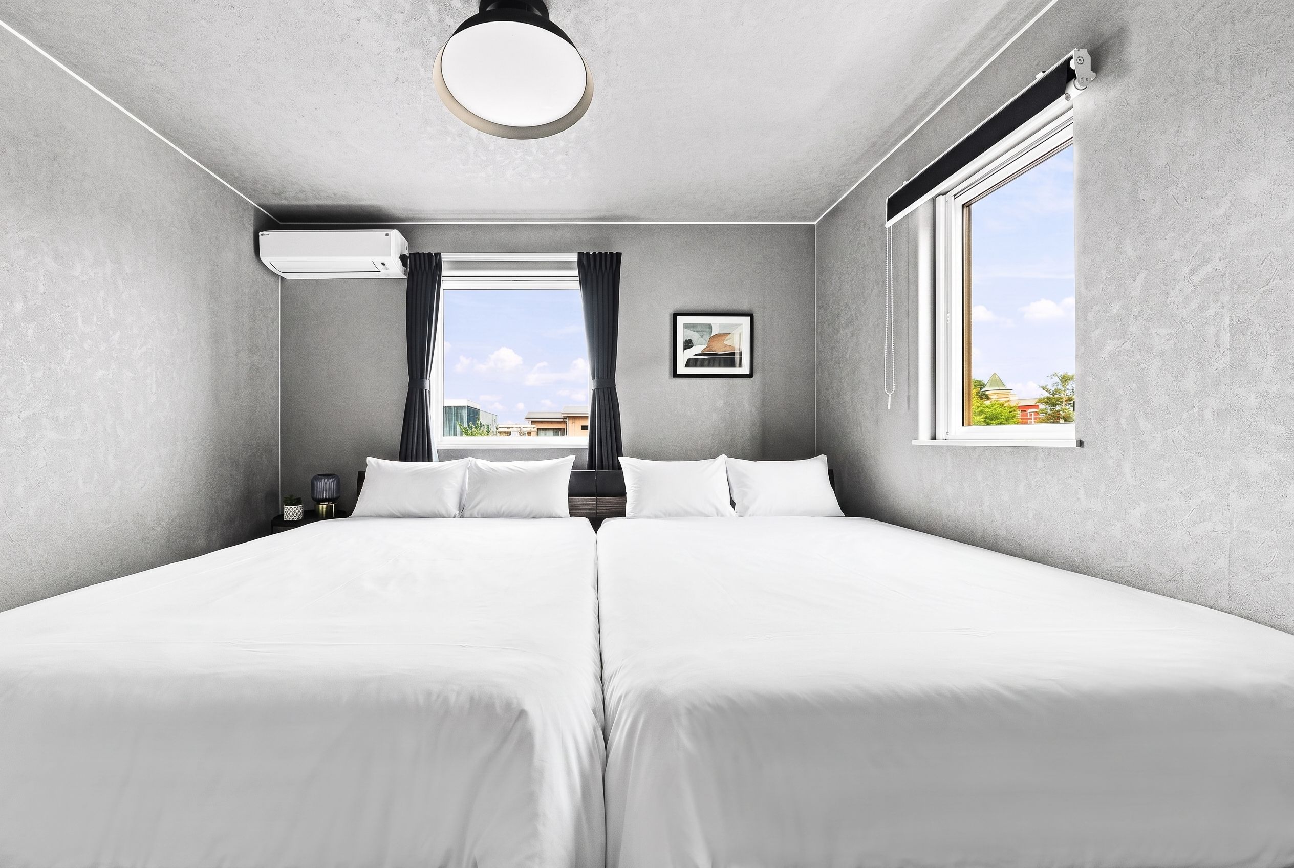 Una camera d'albergo con due letti, una finestra, un condizionatore, un quadro incorniciato e un comodino con una lampada.
