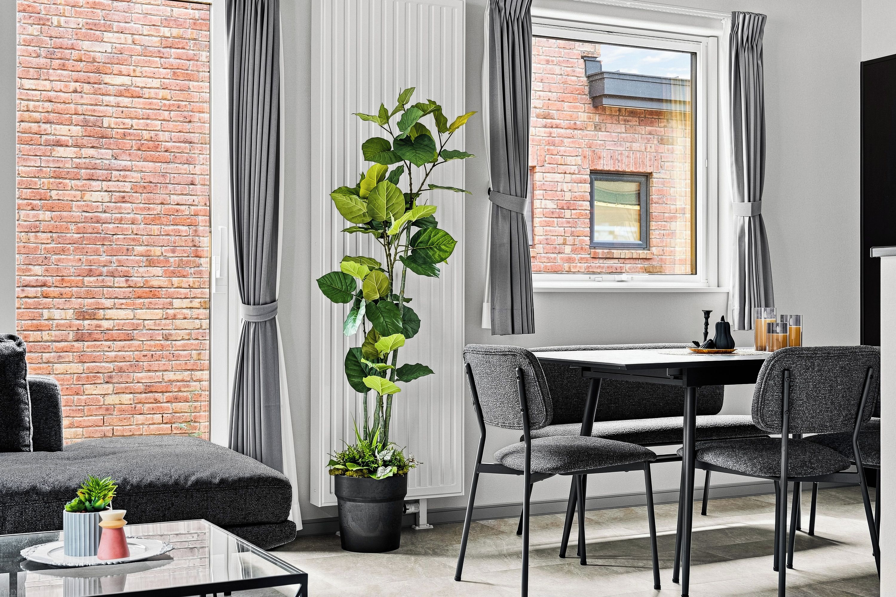 A modern living room with a brick wall, a tall potted plant, grey curtains, a dining table with chairs, and a flat-screen TV.