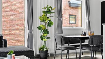 Una sala de estar moderna con una pared de ladrillo, una planta alta en maceta, cortinas grises, una mesa de comedor con sillas y un televisor de pantalla plana.