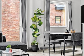 Un saló modern amb una paret de maó, una planta alta en test, cortines grises, una taula de menjador amb cadires i un televisió de pantalla plana.