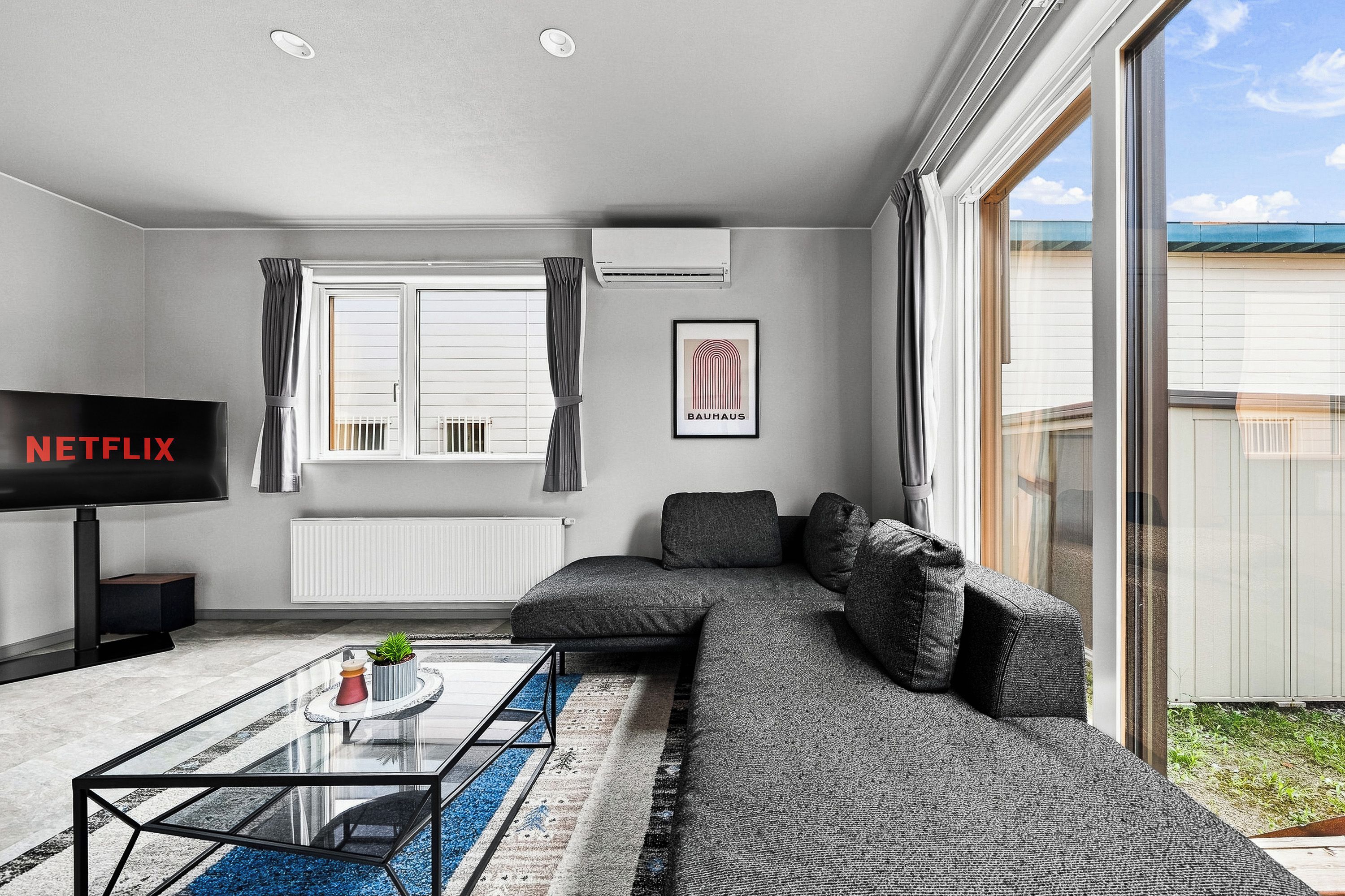 A modern living room with a sectional sofa, a glass coffee table, a flat-screen TV, and a framed Bauhaus logo on the wall.