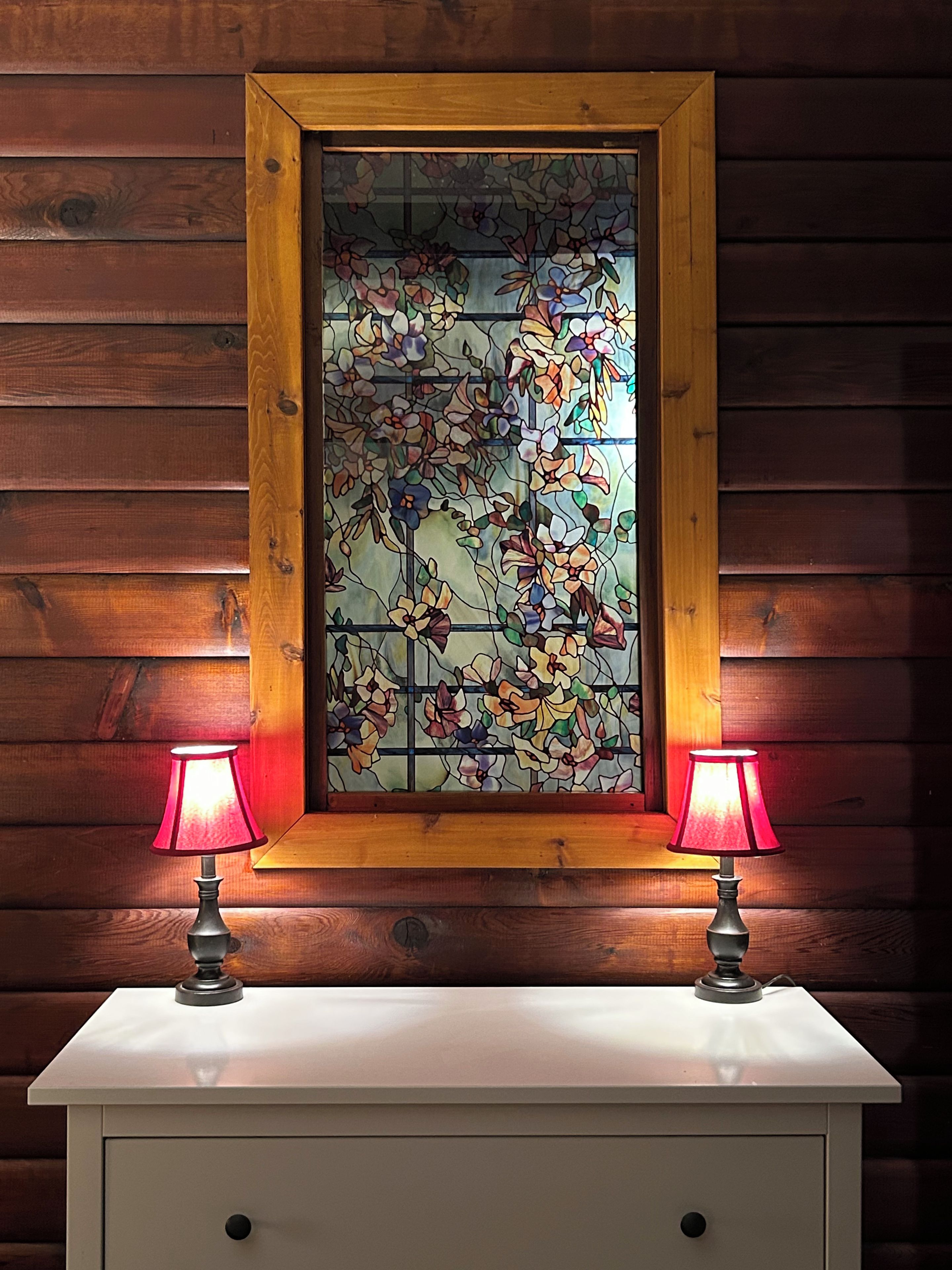 A wooden-framed stained glass window with floral patterns, flanked by two table lamps on a white cabinet.