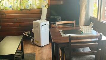 Interior de una cabaña de madera con una mesa de comedor, sillas, un calentador blanco pequeño y una ventana con cortinas.
