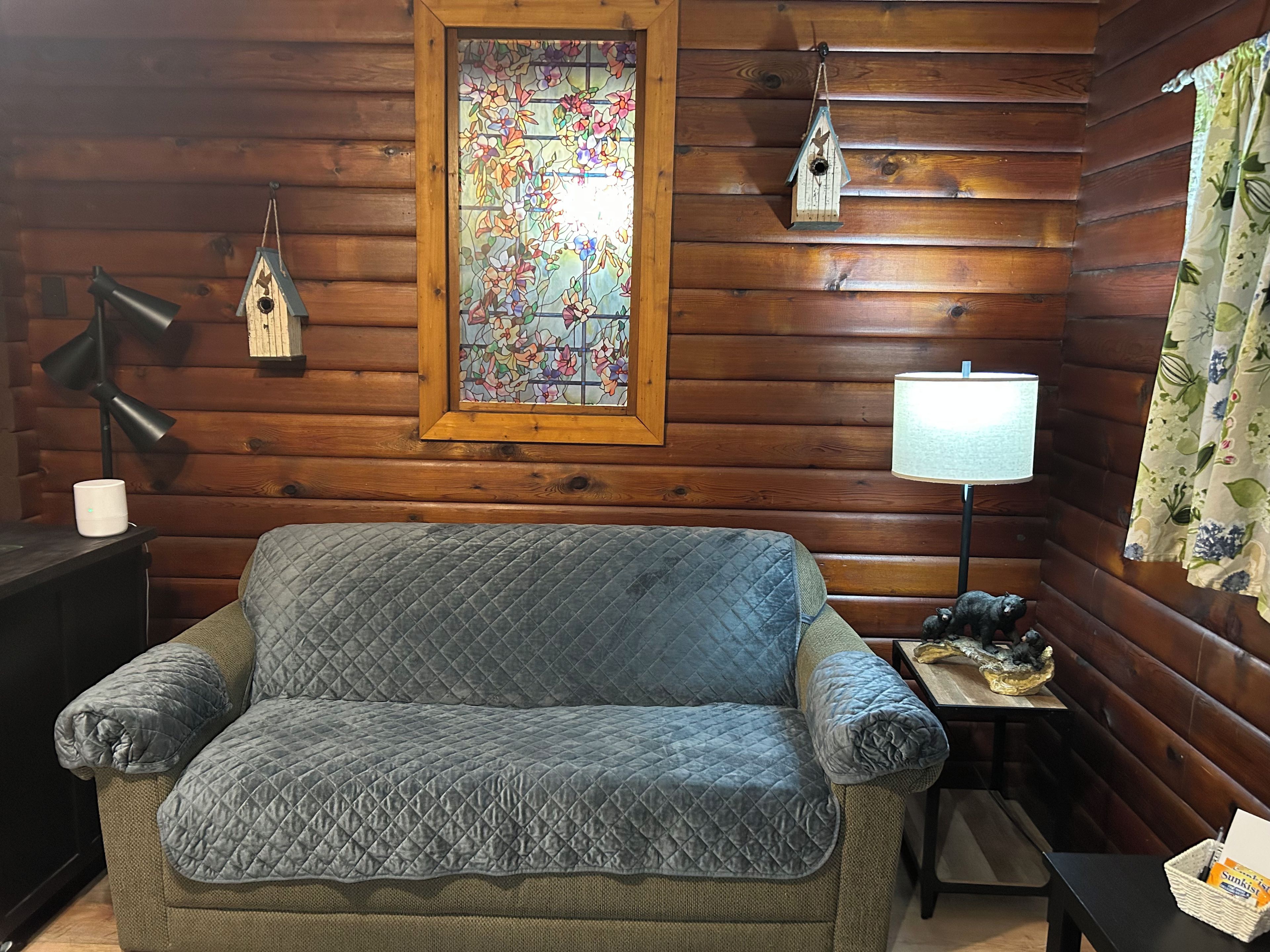 A cozy room with a quilted sofa, a wooden wall, a stained glass window, a lamp, and a small table with a basket.
