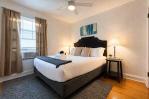 A hotel room with a bed, two bedside lamps, a ceiling fan, a window with curtains, and a painting on the wall. - Lakeview 1BR Apt Near Wrigley & Lake Michigan (Chicago)