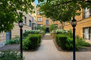 A courtyard with a pathway leading to a building entrance, surrounded by greenery and two street lamps. - Lakeview 1BR Apt Near Wrigley & Lake Michigan (Chicago)